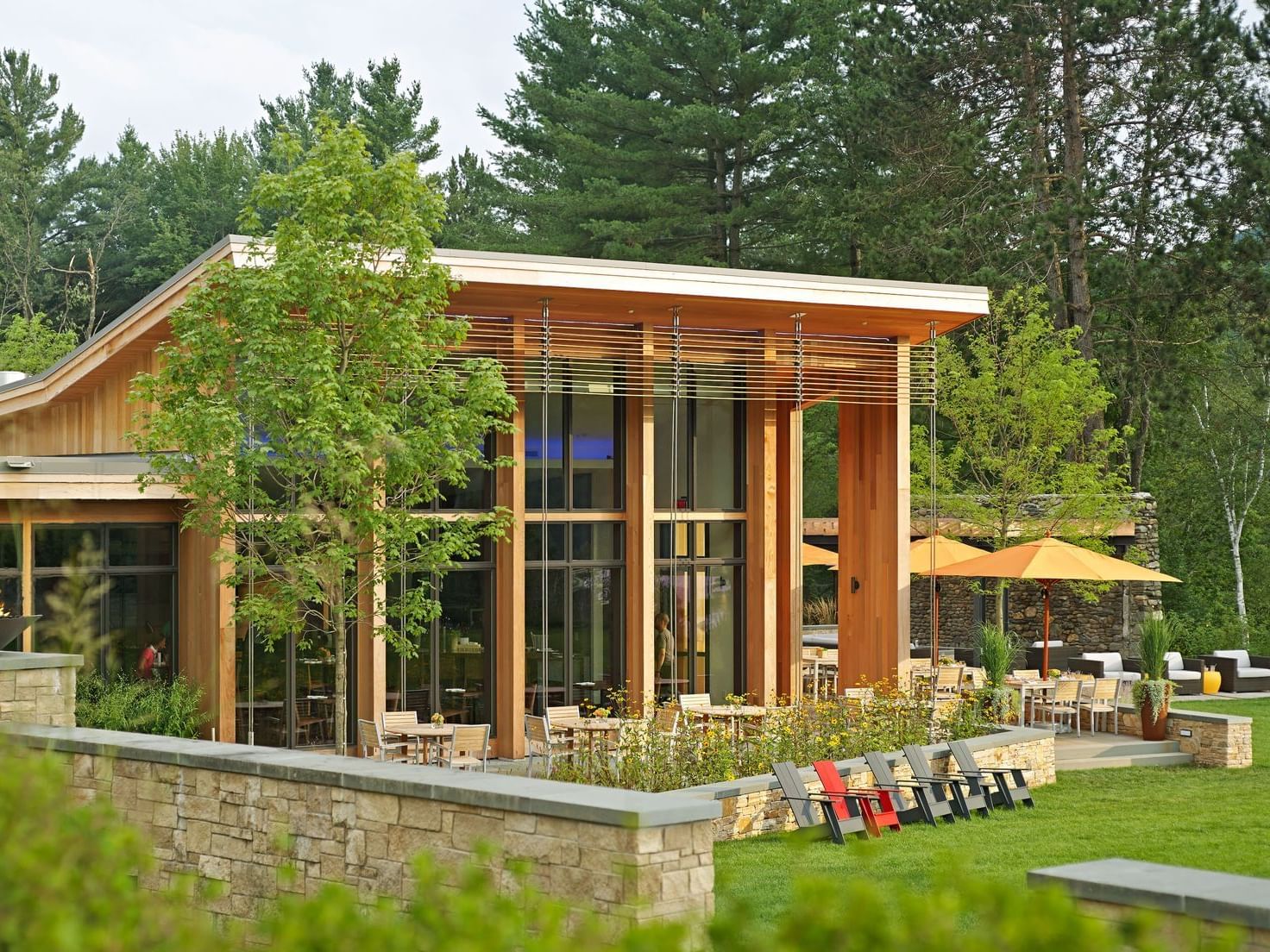 Modern wooden building with patio, tables, chairs, umbrellas, and lush greenery in a forested area.