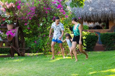 Family of three running in garden with flowers and hut in background.