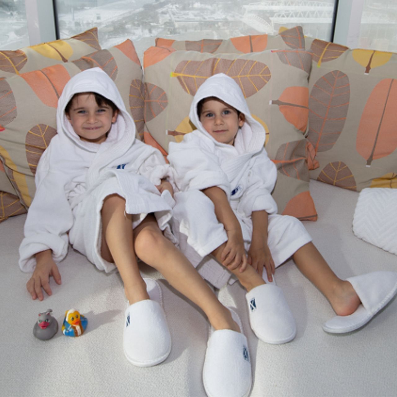 Two kids in white bathrobes and slippers sit on a couch with leaf-patterned pillows