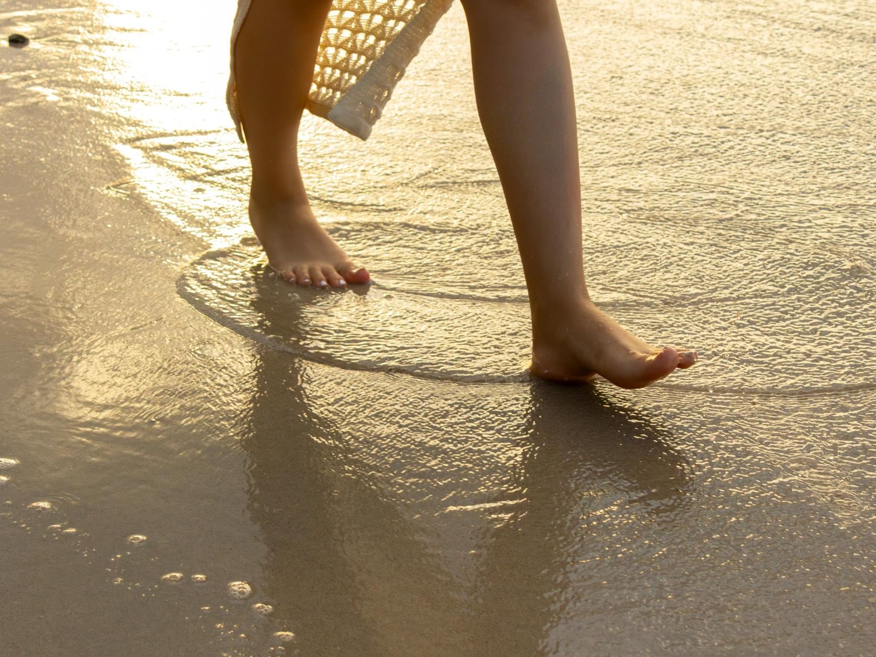 Pies descalzos en la playa con la puesta de sol reflejada en el agua.