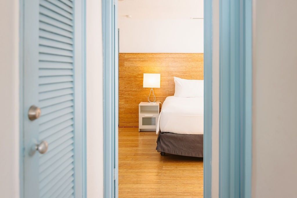 A bed with white linens and a gray bedskirt is visible through an open doorway, with a nightstand and lamp beside it.