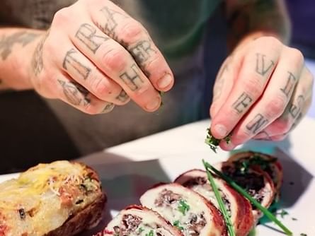 Tattooed hands garnishing sliced meat roulade with fresh herbs alongside a golden baked potato at Jakes Downtown