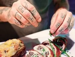 Tattooed hands garnishing sliced meat roulade with fresh herbs alongside a golden baked potato at Jakes Downtown