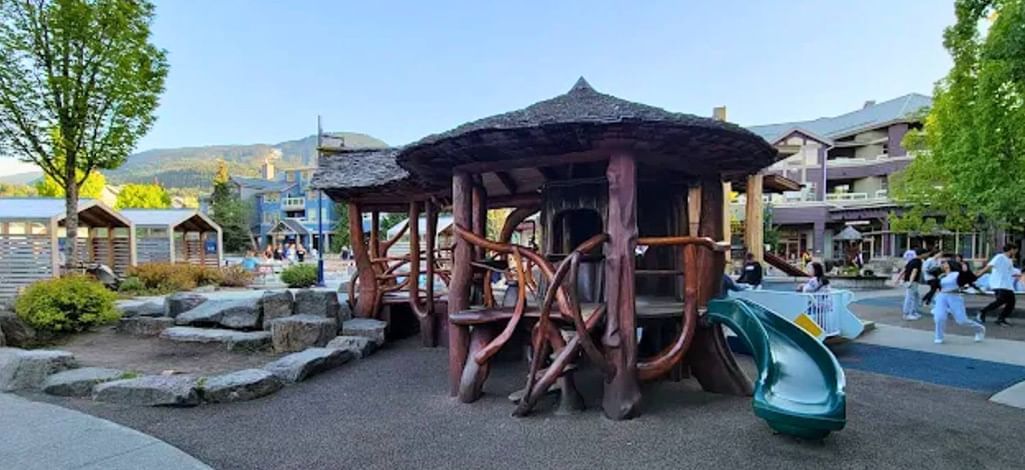 Wooden play structure and rock features at Whistler Olympic Plaza playground with families and nearby buildings in the background.