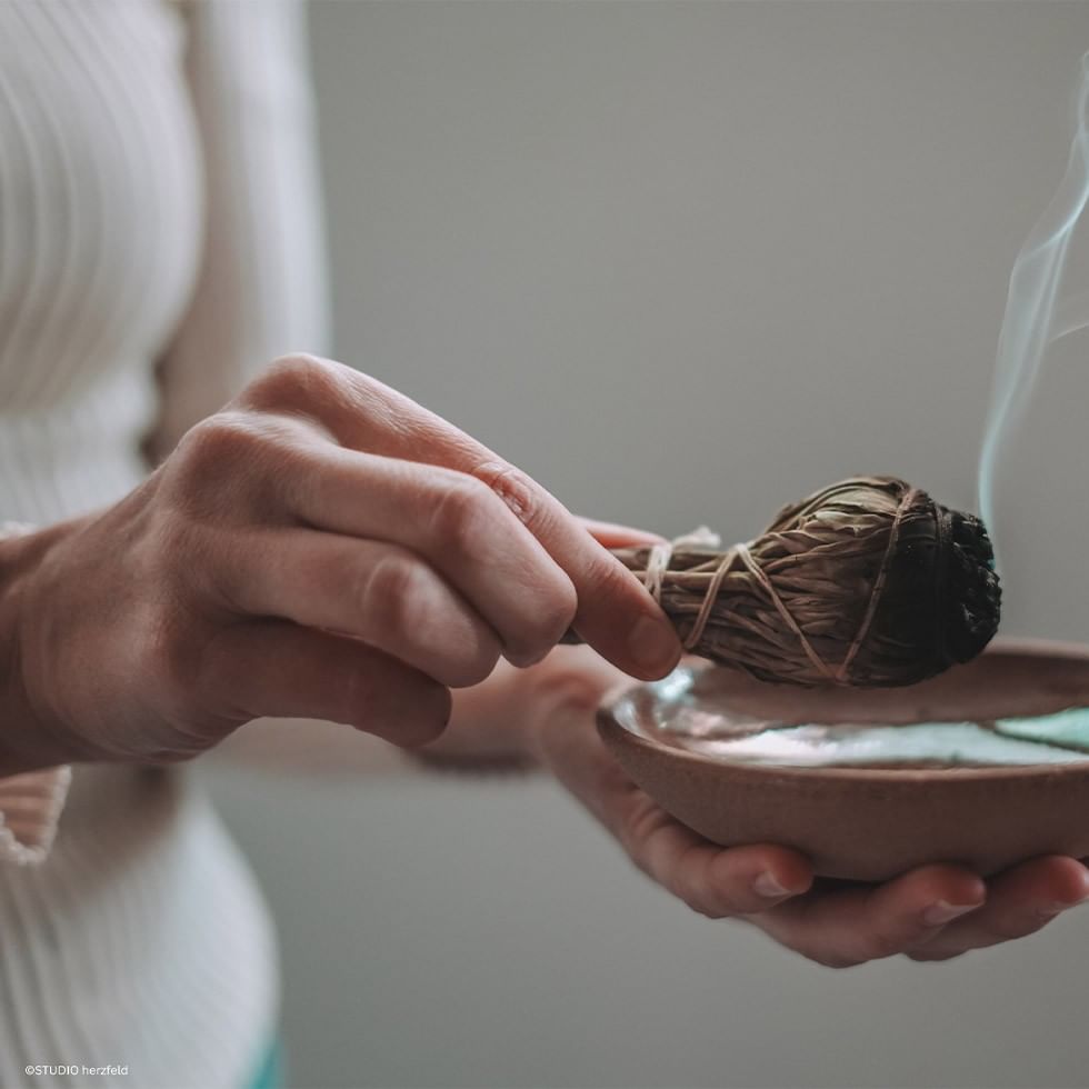 Mani che tengono un fascio fumante sopra una ciotola d'acqua per lo Speciale Yoga Samhain con STUDIO herzfeld