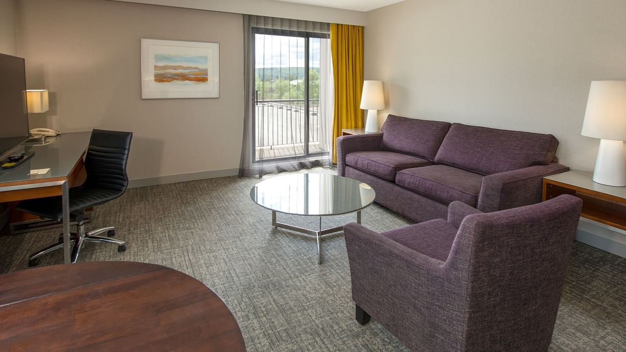 A hotel room with a desk, armchair, couch, work table, and balcony.