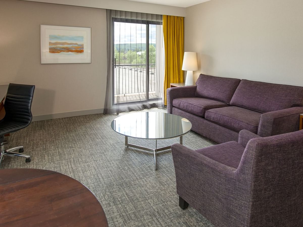 A hotel room with a work desk, armchair, round glass table, and balcony view.