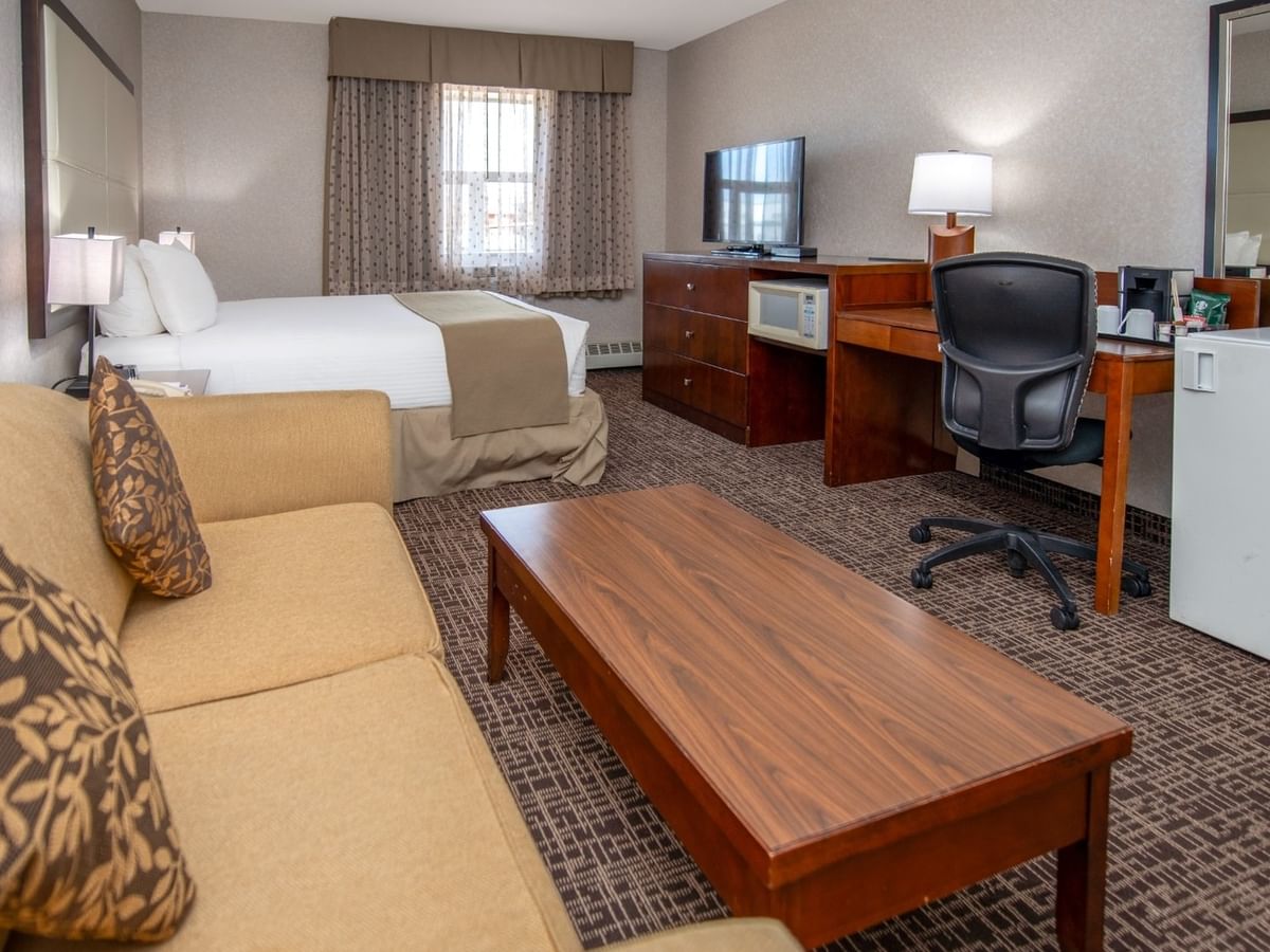 A hotel room with a couch, coffee table, bed, desk, chair, TV, and mini fridge.