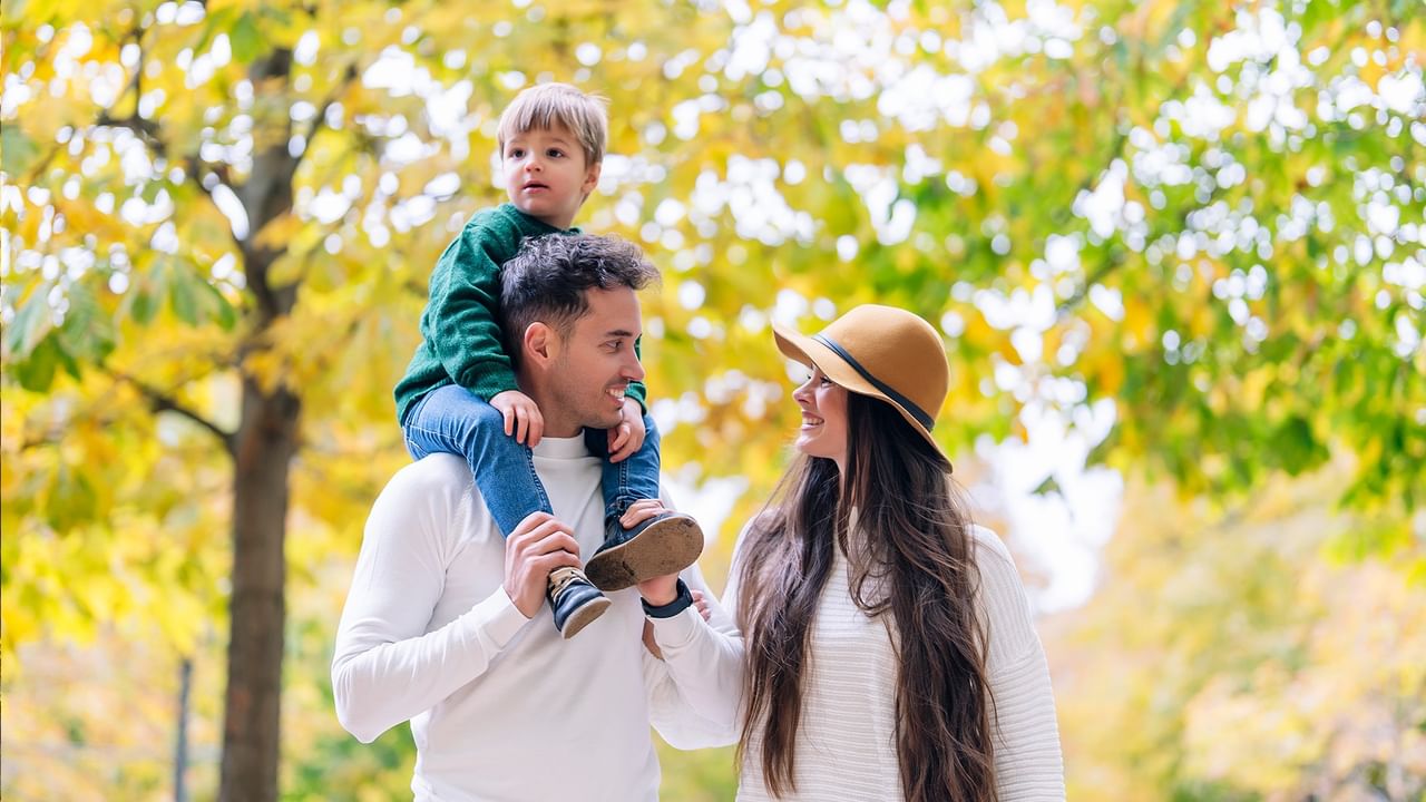 A man carries a child on his shoulders while a woman smiles nearby, all under autumn trees.