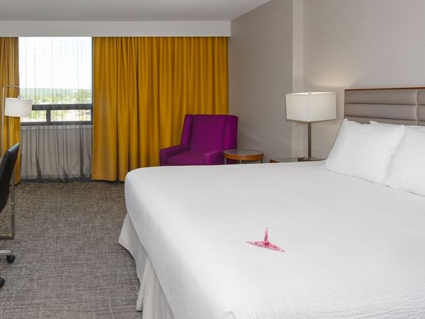 Hotel room with a king bed, desk, purple chair, curtains, and a window.