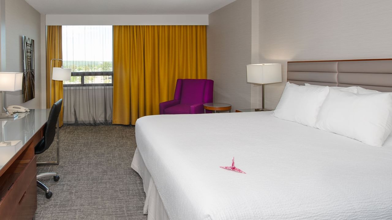 Hotel room with a king bed, desk, purple chair, curtains, and a window.