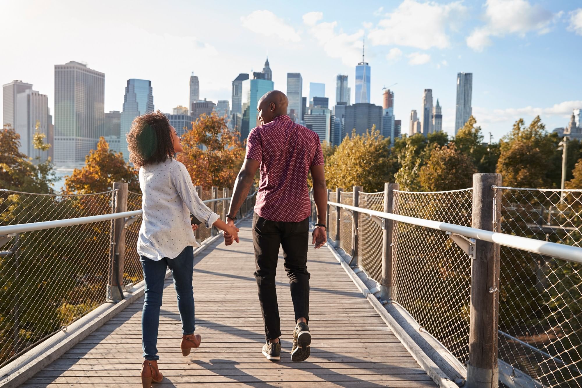 Couple holding hands walking on a wooden bridge with a city skyline in the background at Warwick New York