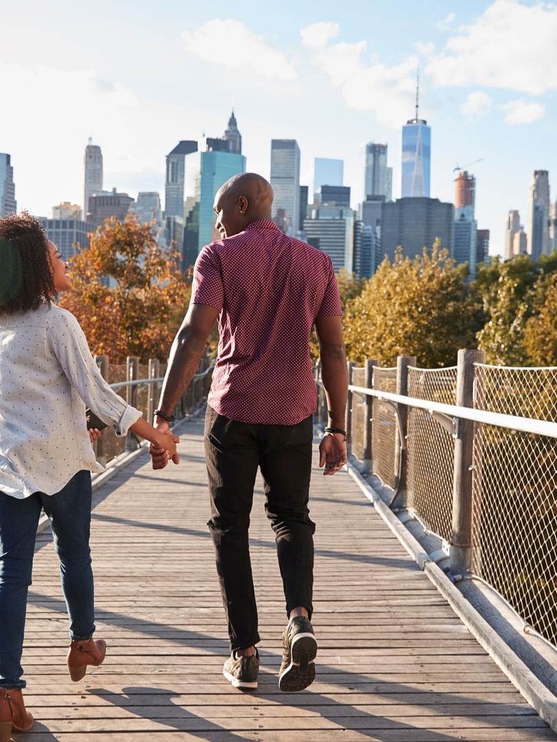 Couple holding hands on a wooden bridge by metal railings, surrounded by fall foliage near Warwick New York