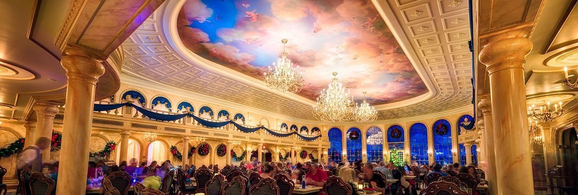A large dining room with a painted ceiling, chandeliers, and many people sitting at tables.
