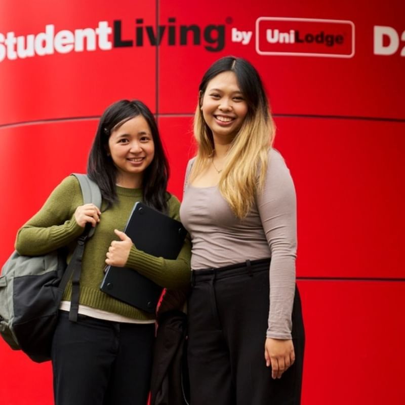 Two smiling students with backpacks standing in front of a bright red StudentLiving by UniLodge branded wall.