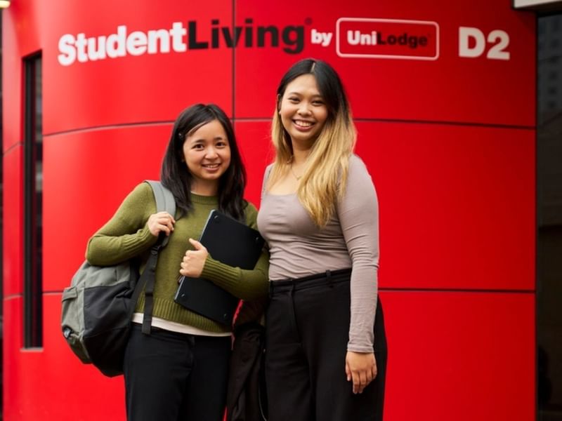 Two smiling students with backpacks standing in front of a bright red StudentLiving by UniLodge branded wall.
