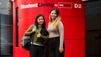 Two smiling students with backpacks standing in front of a bright red StudentLiving by UniLodge branded wall.