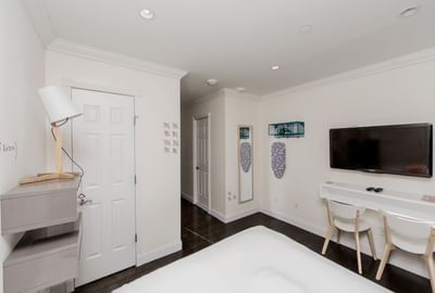 East Village Hotel bedroom with bed, desk, chairs, TV, lamp, and door leading to another room.