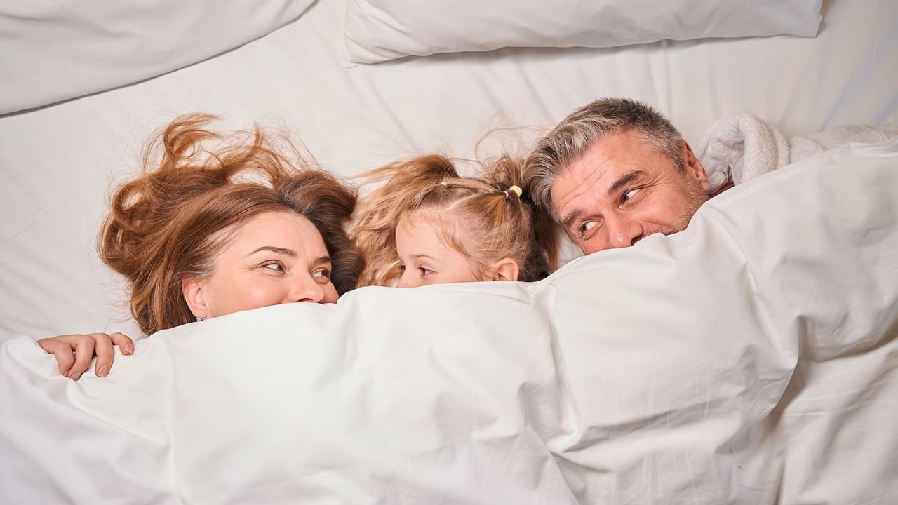 Family of three smiling under the blankets on a hotel bed.