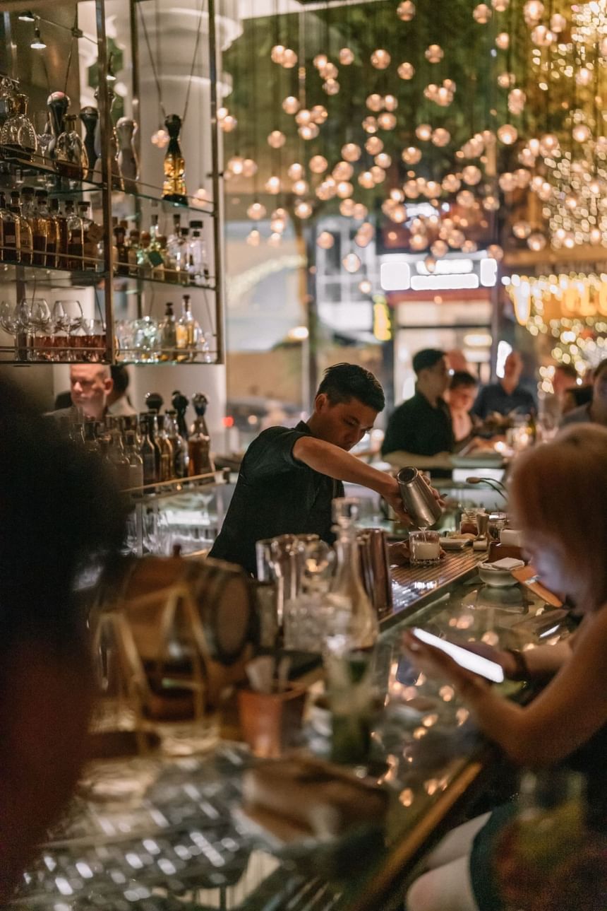 Bartender pouring a drink at a lit bar, surrounded by guests and shelves of bottles in 2 Lam Son at Park Hyatt Saigon