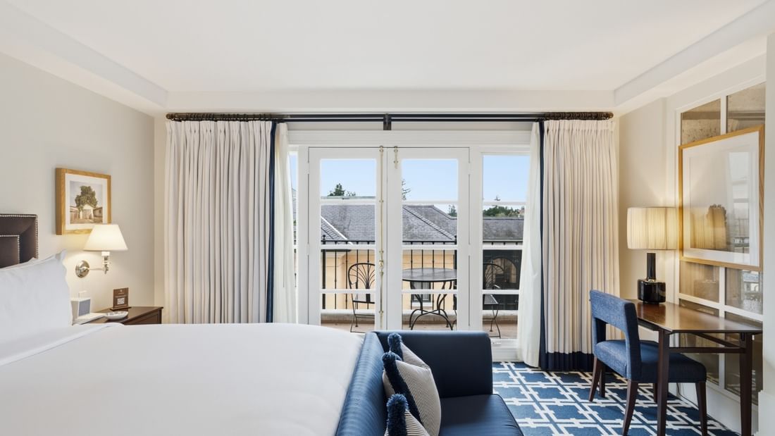 Deluxe King room with white bedding, blue accent pillows, balcony doors, and geometric carpet at el PRADO Hotel