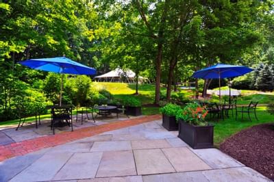 Outdoor patio with blue umbrellas and black iron tables on stone paving between lush green trees at Avon Old Farms Hotel