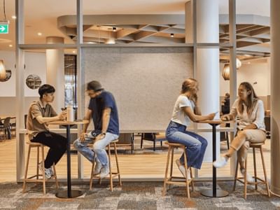 Four people sit at tables in a modern office space with glass walls and wooden floors.