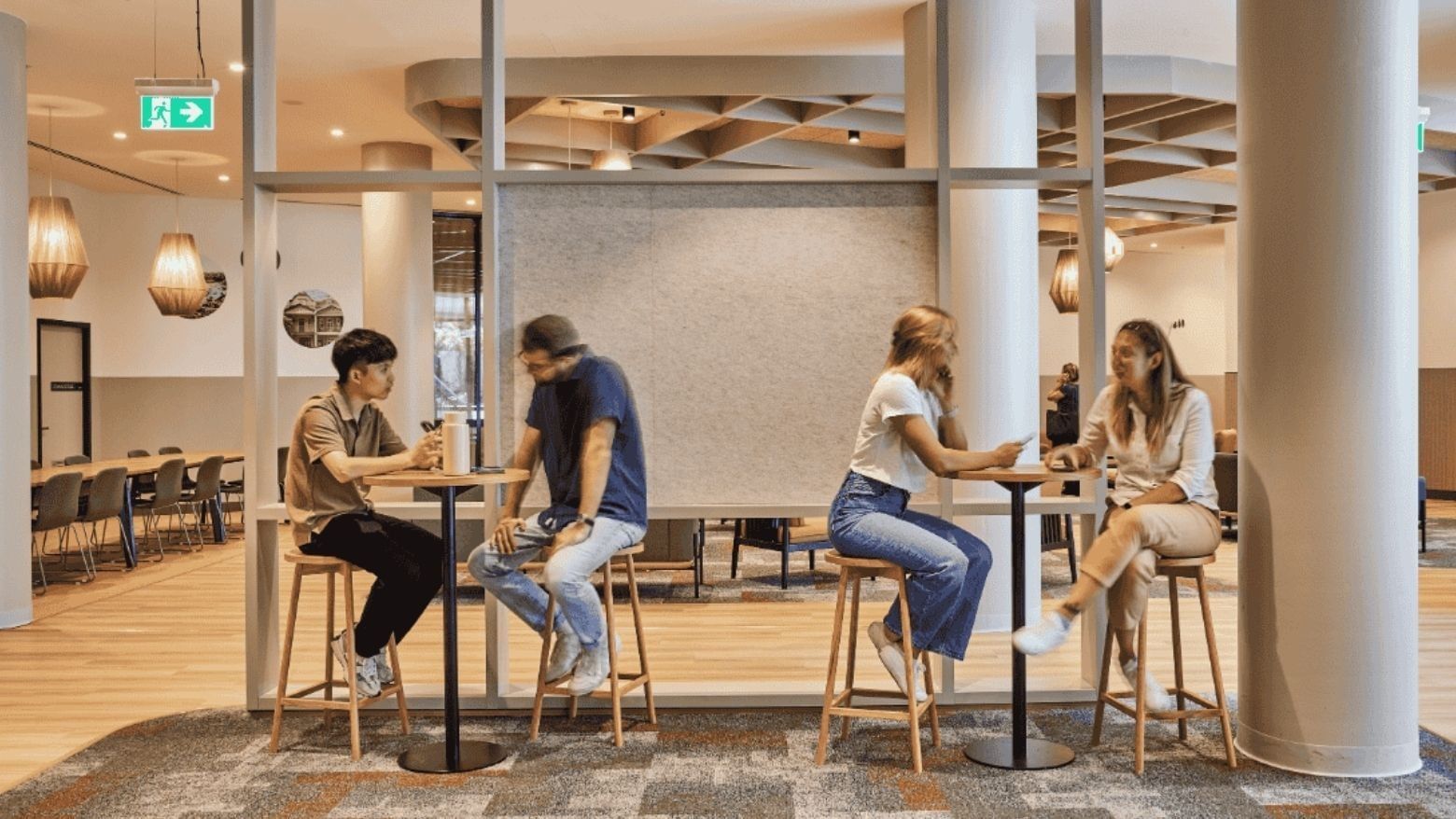 Four people sit at tables in a modern office space with glass walls and wooden floors.