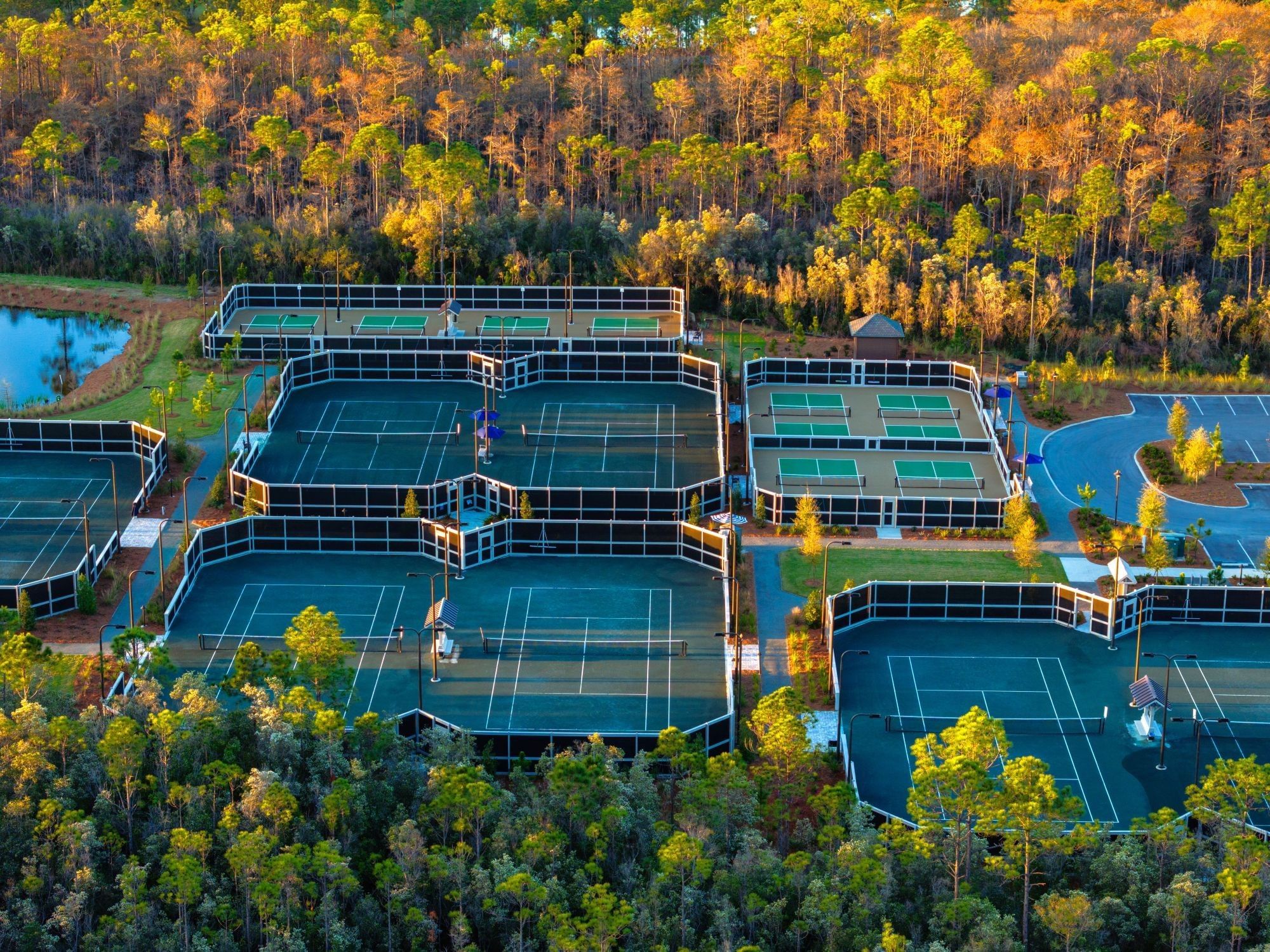 Aerial view of a tennis complex with multiple courts, surrounded by trees and a pond.