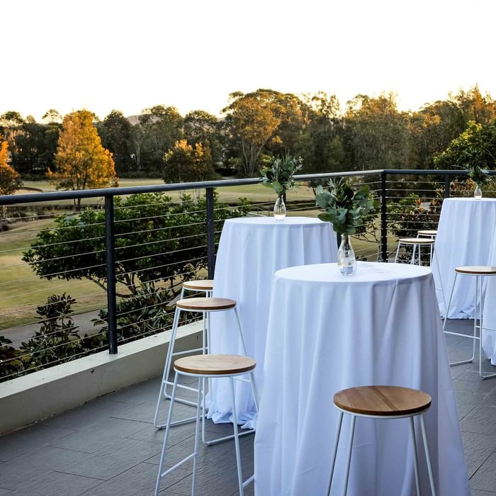 Four round tables with white cloths and greenery in vases on a balcony overlooking a park.