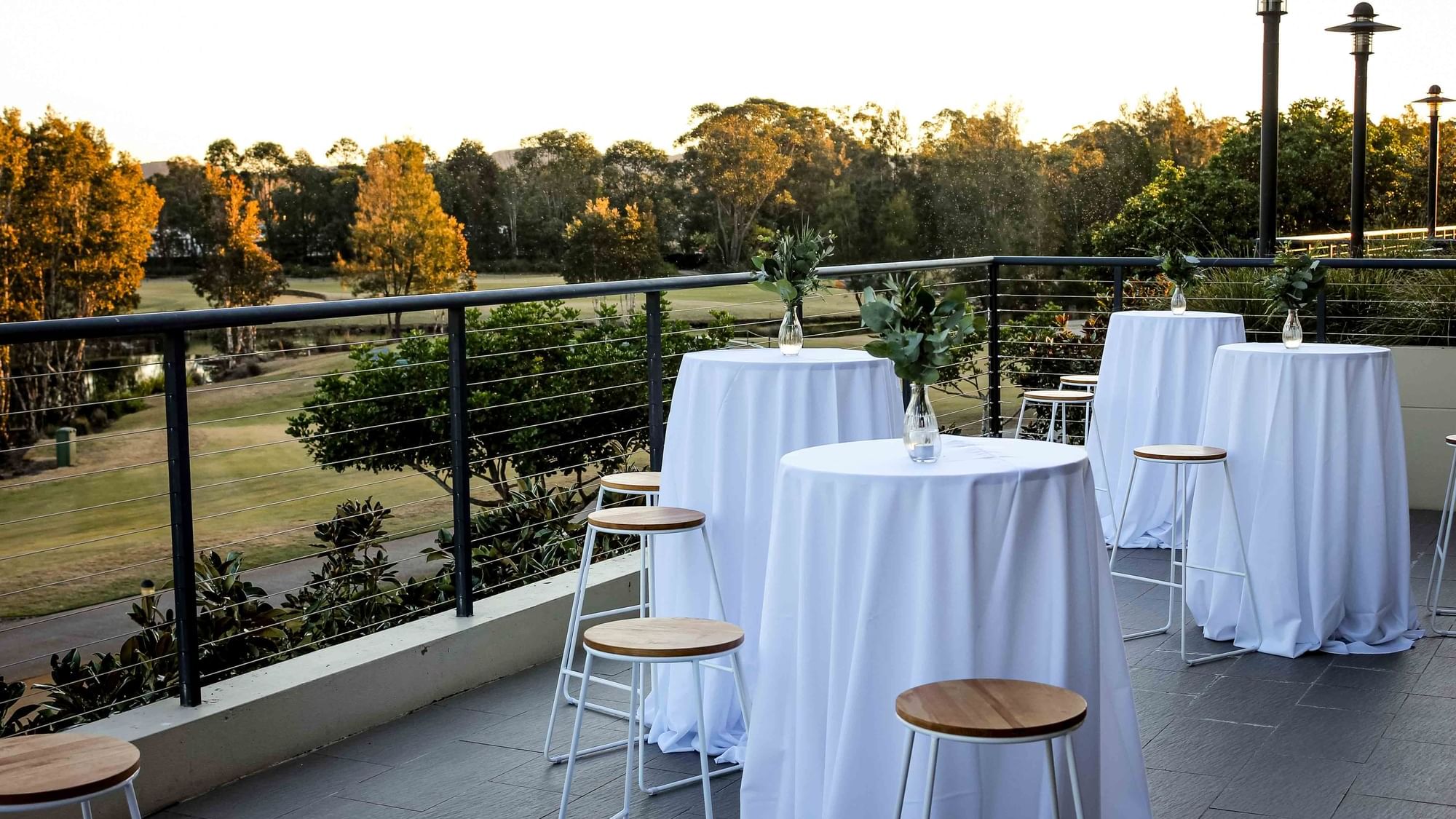 Four round tables with white cloths and greenery in vases on a balcony overlooking a park.