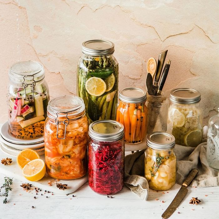 Several jars filled with pickled vegetables and herbs, including lemon slices, on a white surface with kitchen tools.