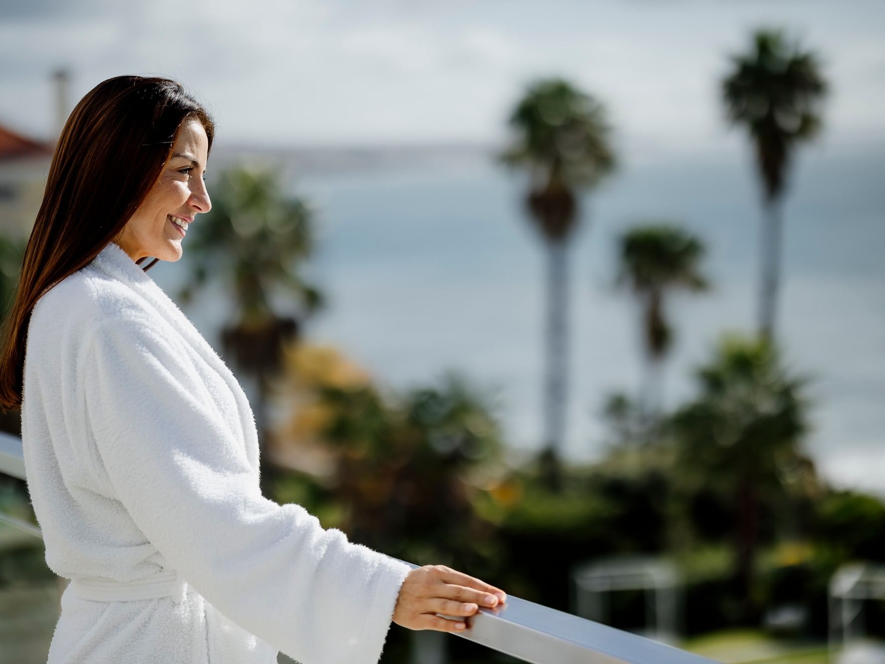 Woman in white bathrobe smiling on balcony overlooking palm trees and ocean view.