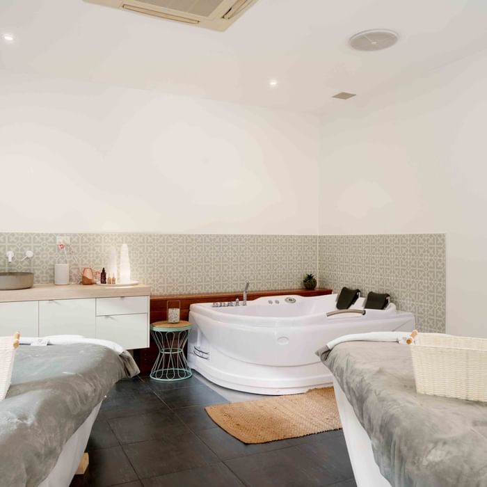 Two beds with wicker baskets, a large bathtub, and a patterned wall in a white room.