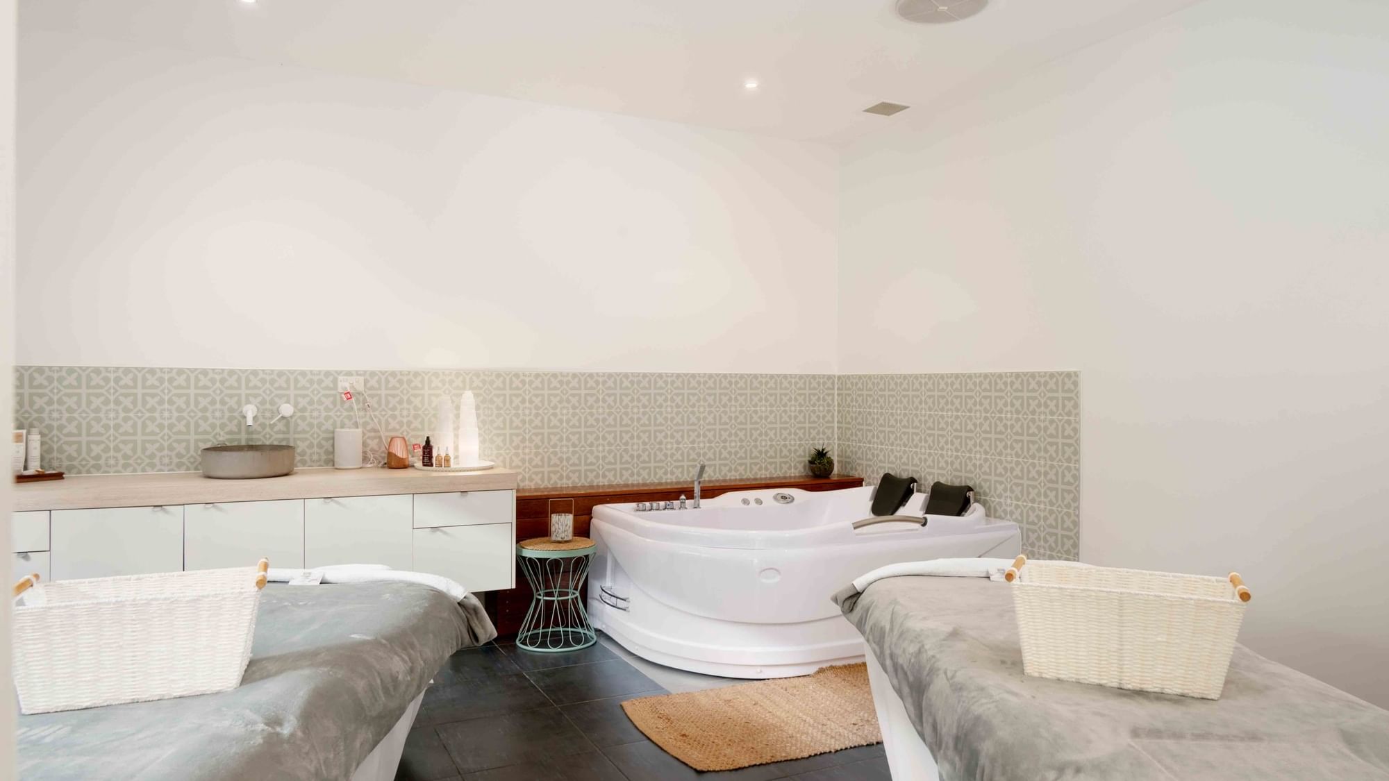 Two beds with wicker baskets, a large bathtub, and a patterned wall in a white room.