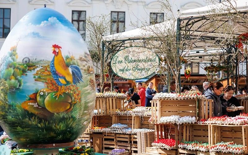 Wiener Ostermarkt mit großem kunstvoll bemaltem Osterei und Marktständen im Hintergrund.