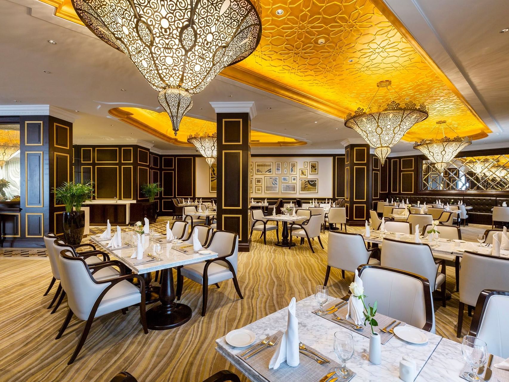 Elegant restaurant with white tables, chairs, decorative chandeliers, and ornate ceiling designs.
