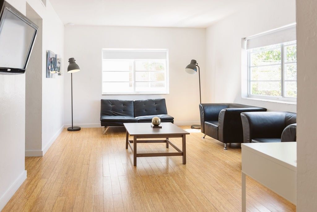 A bright living room with a black leather sofa and armchair, a wooden coffee table, and a TV on the wall.