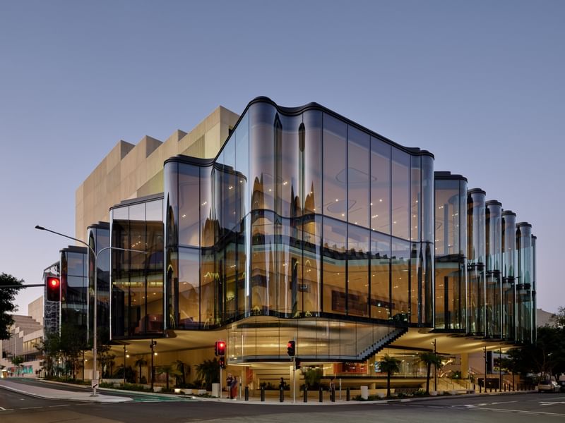 Modern curved glass facade of QPAC illuminated at dusk with reflective architectural panels and street-level entrance.