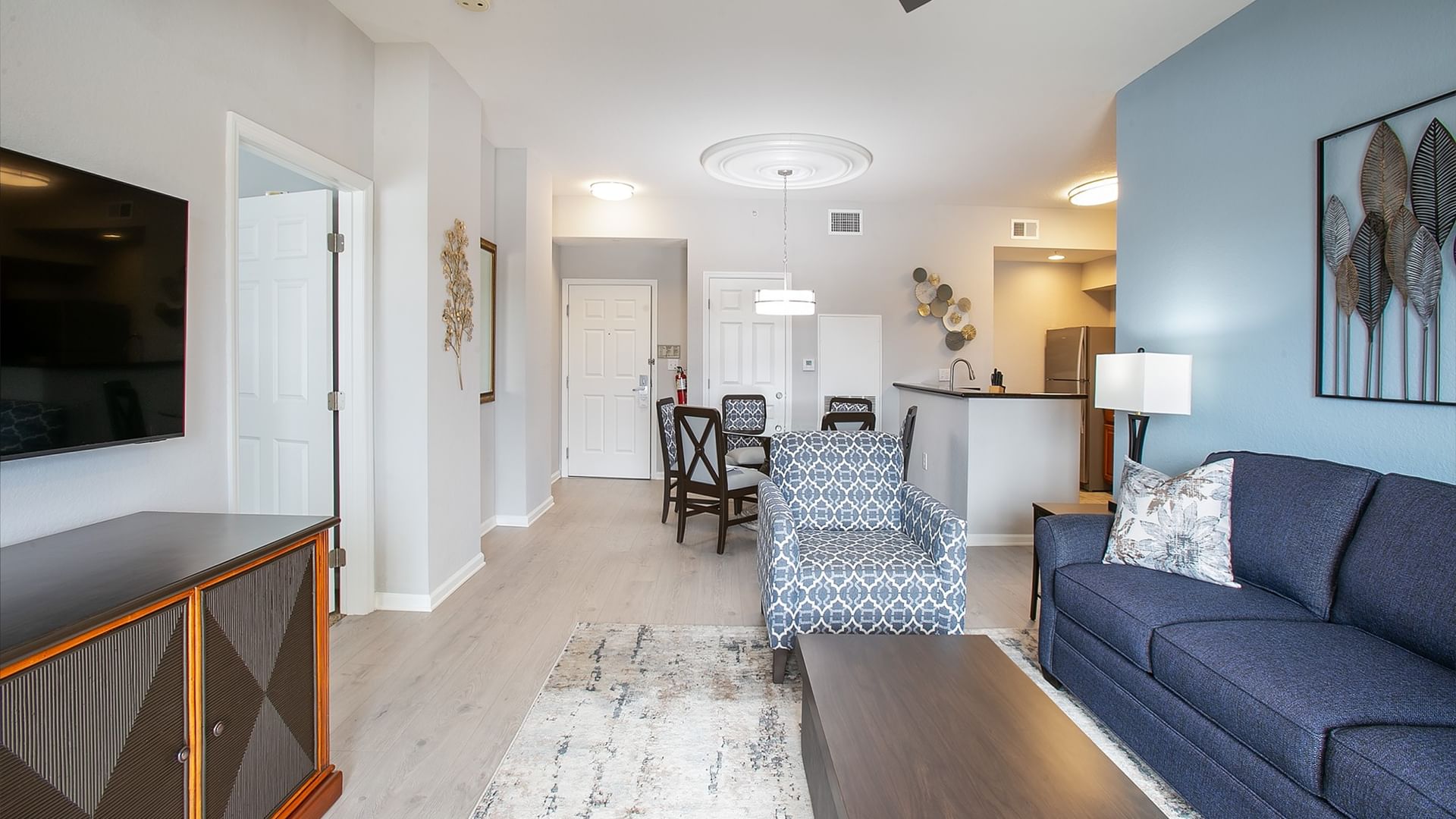 A wide view of a living room with a blue sofa, coffee table, TV, dining table, and kitchenette.