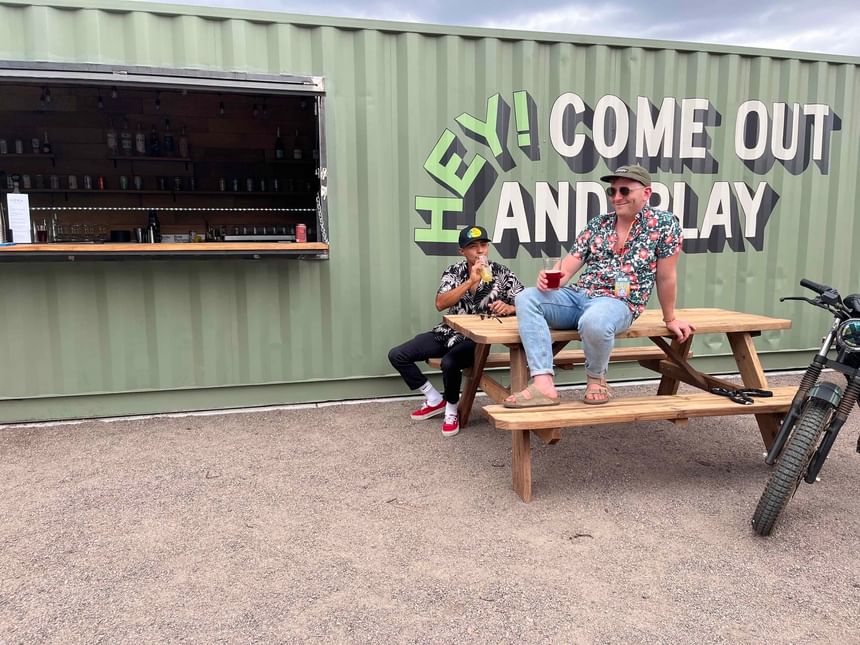 Two men having beers sitting on a wooden bench outdoors at Kinship Landing