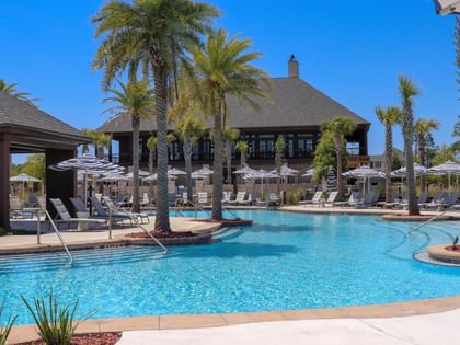 Outdoor pool area with palm trees, lounge chairs, and a large pavilion building under a clear blue sky.