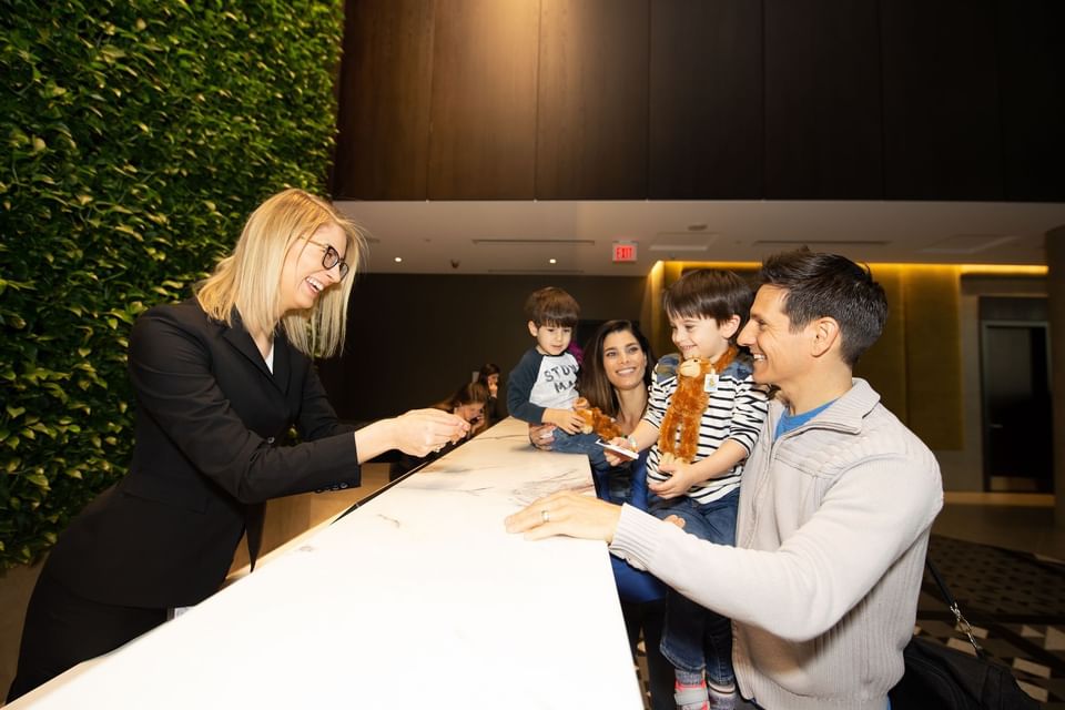 Smiling receptionist greeting family with young children at white marble front desk beside green wall at Hotel X Toronto