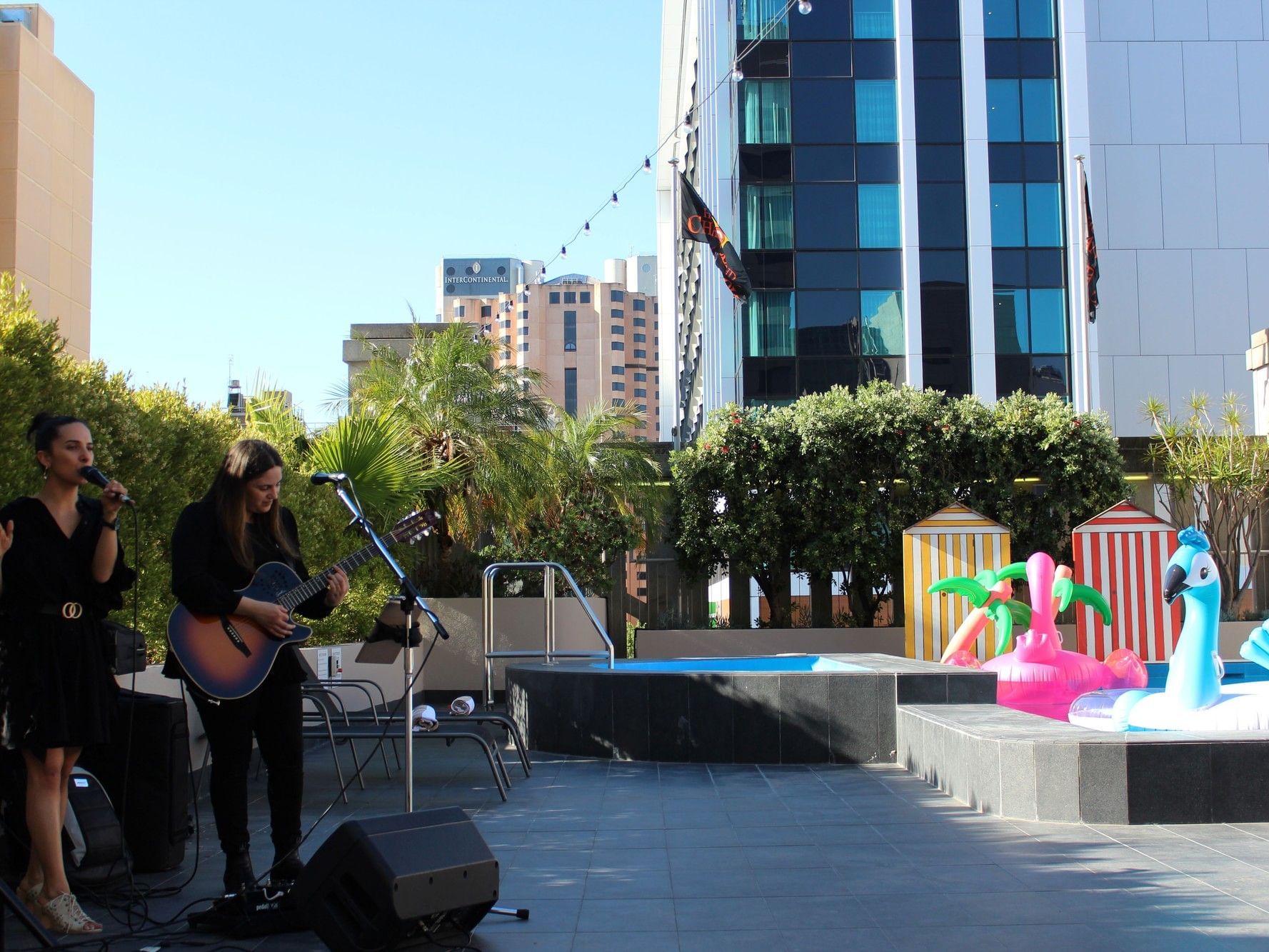 Two women performing poolside with a guitar and microphone, inflatable beach toys nearby.
