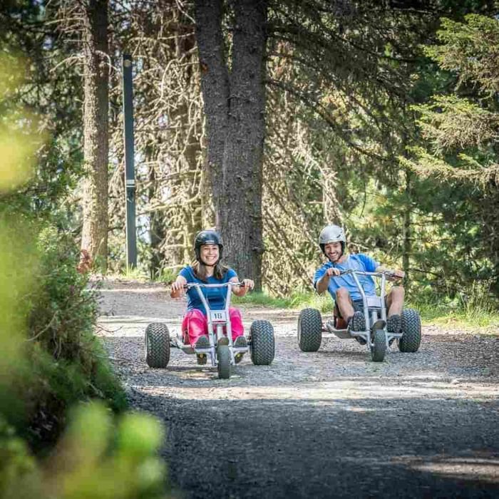 Due persone guidano go-kart su una strada sterrata nel bosco con alberi alti e luce solare.
