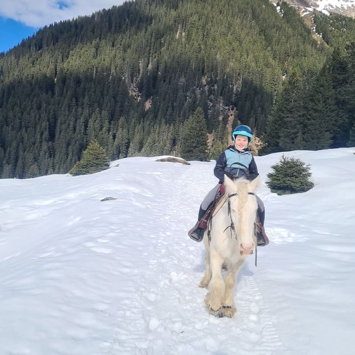 Kind mit Helm reitet ein weißes Pferd im schneebedeckten Wald vor einem Berg.