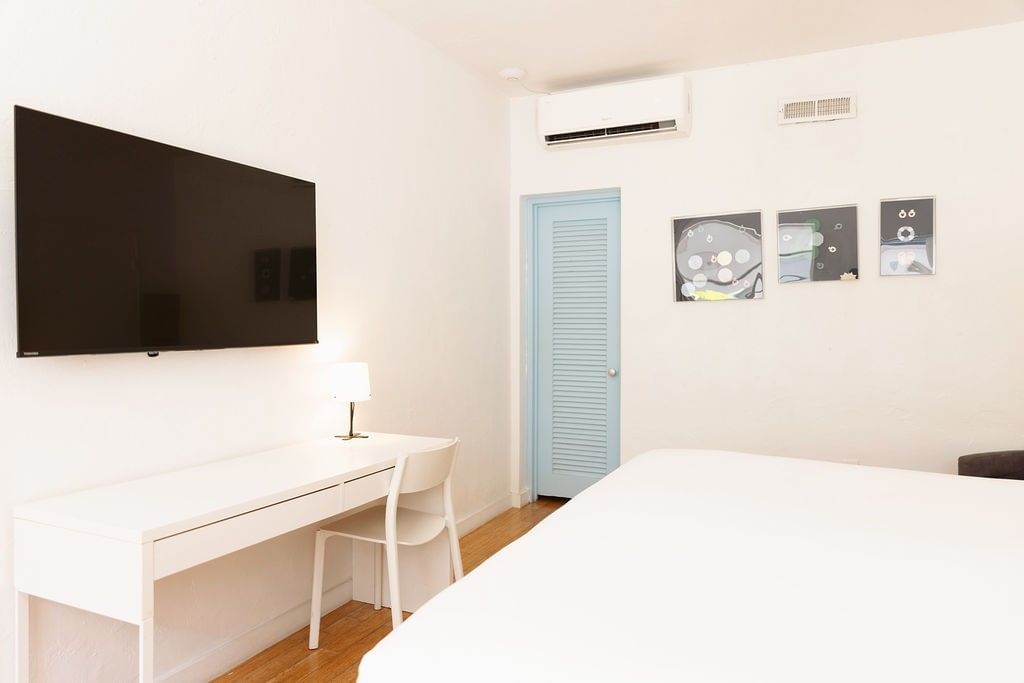 A white hotel room with a bed, desk, chair, lamp, and TV