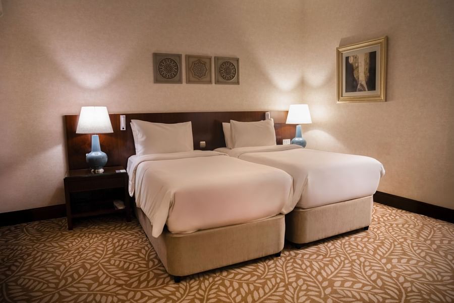 Twin bedroom featuring side lamps with wall art and patterned brown and beige carpet at Saja by Warwick Makkah