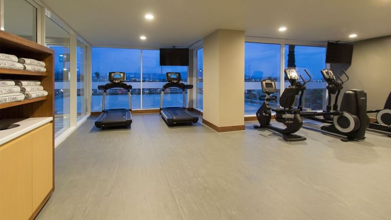 Modern gym with treadmills and ellipticals, large windows showing an evening sky at Fiesta Inn Suites Aeropuerto del Bajío