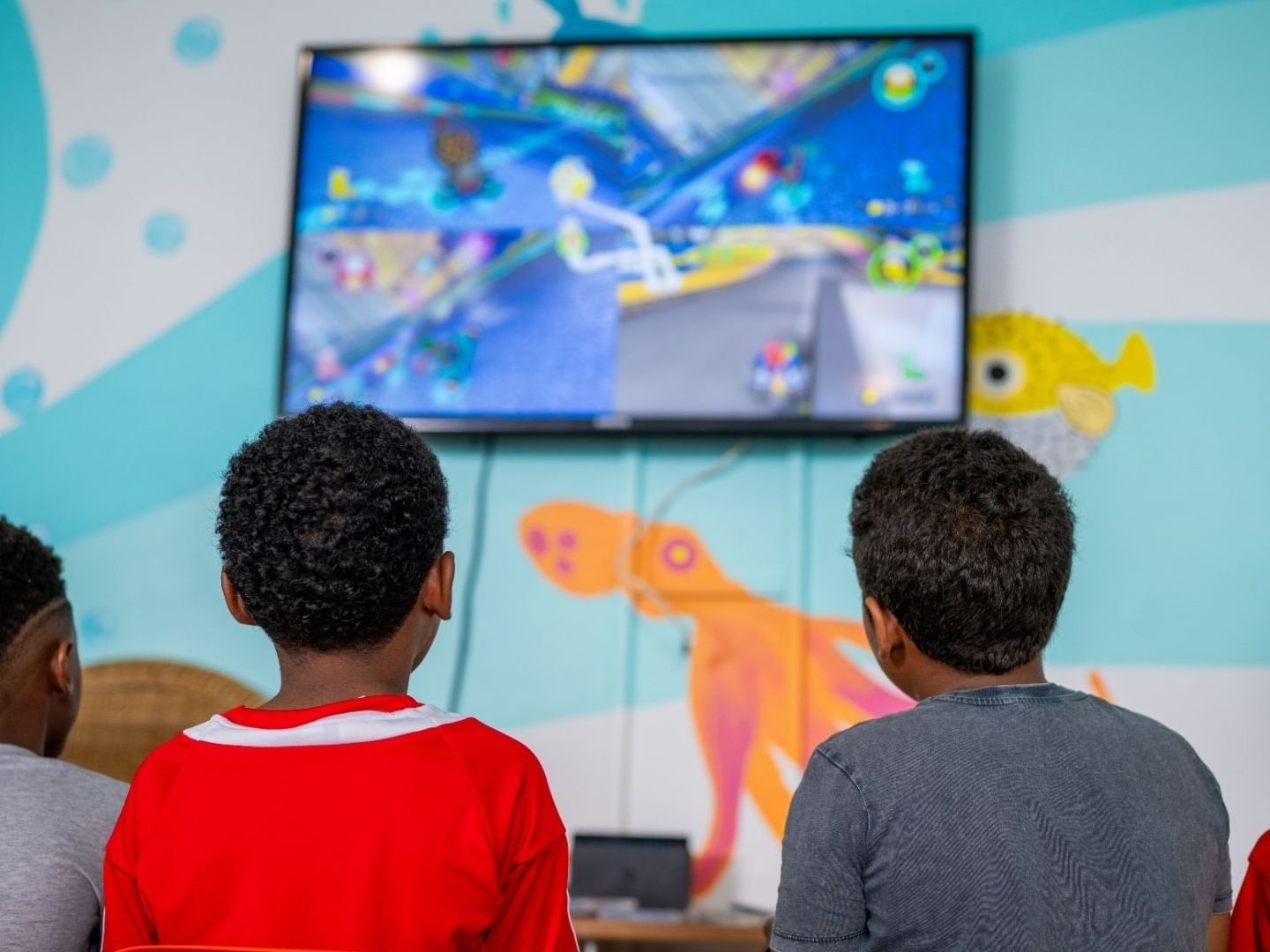 Four children watching a game on a mounted TV in a room with aquatic wall decorations at Savannah Beach Hotel.